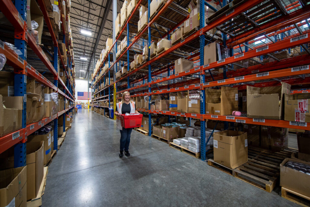 woman in reflective vest carrying red box through an aisle of a warehouse